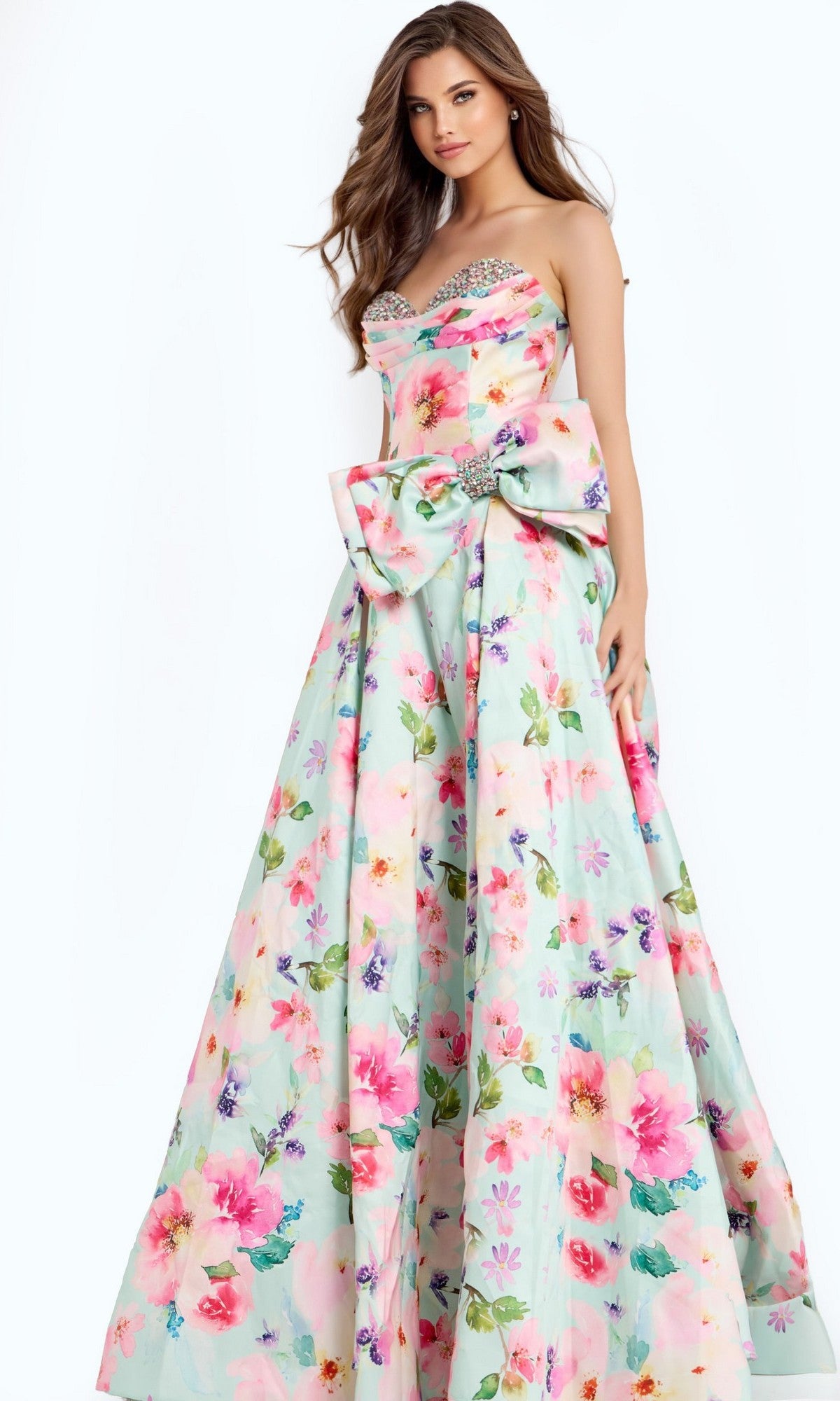 A woman models the Jovani 49197 long prom dress, featuring a strapless floral A-line silhouette with a sweetheart neckline, corset bodice, and large waist bow, set against a plain light background.